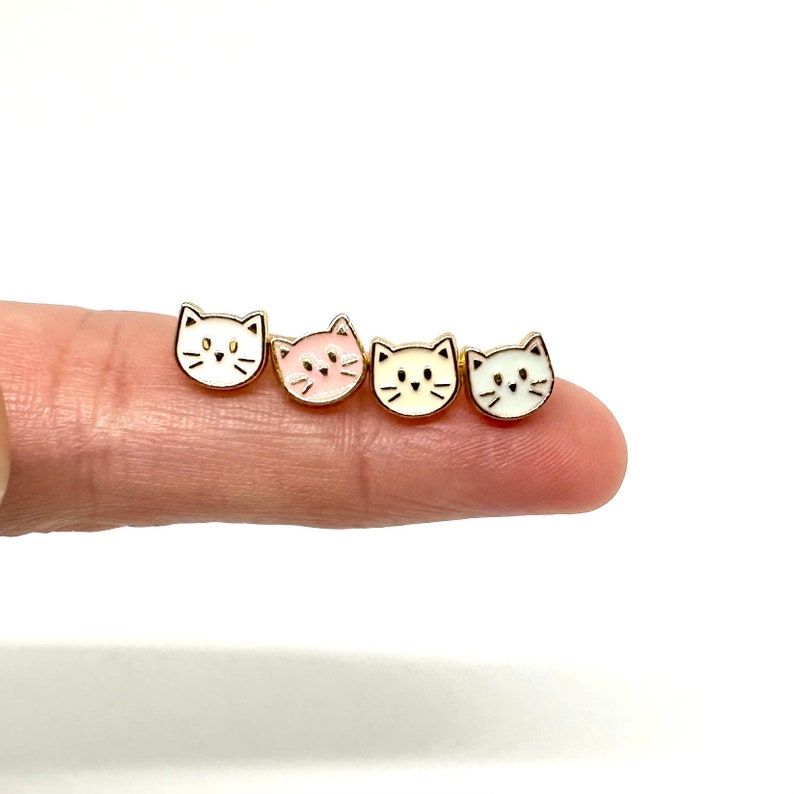 May include: Four gold cat face earrings with white, pink, and yellow enamel. The earrings are on a finger.