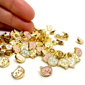 May include: A large pile of gold-colored metal buttons with a white enamel cat face design. The buttons are in various shades of pink, white, and cream.