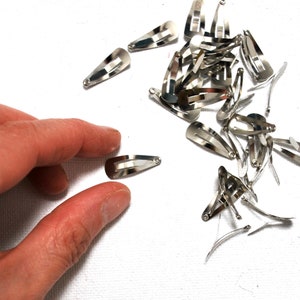 Op de afbeelding: Een verzameling kleine, zilverkleurige metalen haarclips verspreid op een wit oppervlak. De clips zijn druppelvormig met een veermechanisme. Een hand reikt naar een van de clips.