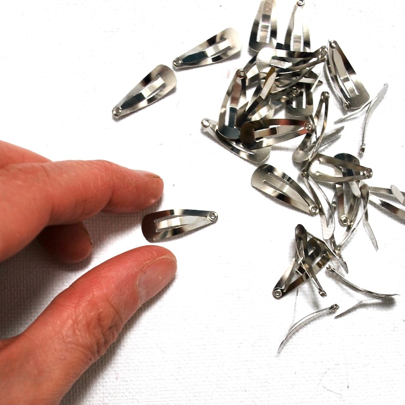 Op de afbeelding: Een verzameling kleine, zilverkleurige metalen haarclips verspreid op een wit oppervlak. De clips zijn druppelvormig met een veermechanisme. Een hand reikt naar een van de clips.