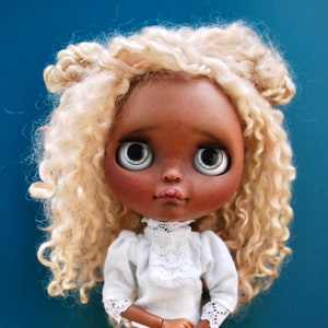 May include: A doll with blonde curly hair and brown skin wearing a white lace blouse. The doll has large blue eyes and a serious expression.