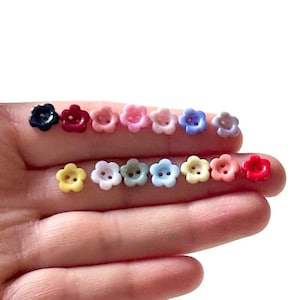 Könnte beinhalten: Eine Sammlung kleiner, blütenförmiger Knöpfe in verschiedenen Farben, darunter Marineblau, Rot, Rosa, Blau, Gelb und Weiß. Jeder Knopf hat ein Vier-Loch-Design zum Nähen. Die Knöpfe werden in einer Hand vor weißem Hintergrund gehalten.