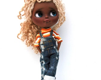 May include: A doll with blonde curly hair, brown skin, blue eyes, and a serious expression. She is wearing a striped orange and white shirt and blue denim overalls.