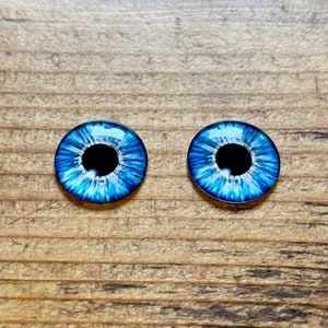 Op de afbeelding: Twee blauwe plastic poppenogen met zwarte pupillen en witte iris. De ogen liggen op een houten oppervlak.