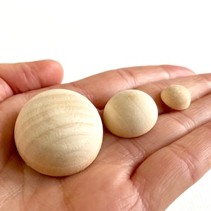 May include: Three wooden spheres of varying sizes, displayed on a hand. The spheres are smooth and light brown in color.