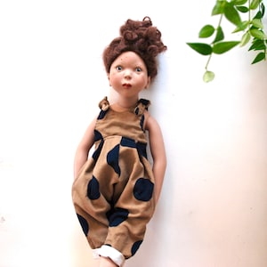 Könnte beinhalten: Eine Puppe mit braunen Locken, die einen braunen und blauen Tupfen-Overall trägt.