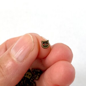 May include: A small, antique brass-colored cat-shaped charm with two eye holes and a rounded head. The charm is held between fingers against a white background. Other similar charms are visible at the bottom.