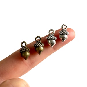 Peut inclure: Quatre breloques en forme de glands en argent et bronze. Les breloques sont présentées sur un doigt, les breloques en argent à gauche et les breloques en bronze à droite.