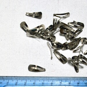 Op de afbeelding: Een verzameling kleine, zilverkleurige metalen haarclips verspreid over een wit oppervlak. De clips zijn ovaal van vorm met een veermechanisme. Een liniaal met centimeter markeringen is zichtbaar in het onderste deel van de afbeelding.