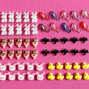 May include: A collection of small, colorful resin charms arranged on a pink surface. The charms include white bunnies with "LOVE" text, fish, bats, brown bears with heart details, pink bunnies, and yellow ducklings. These decorative charms are ideal for crafting.