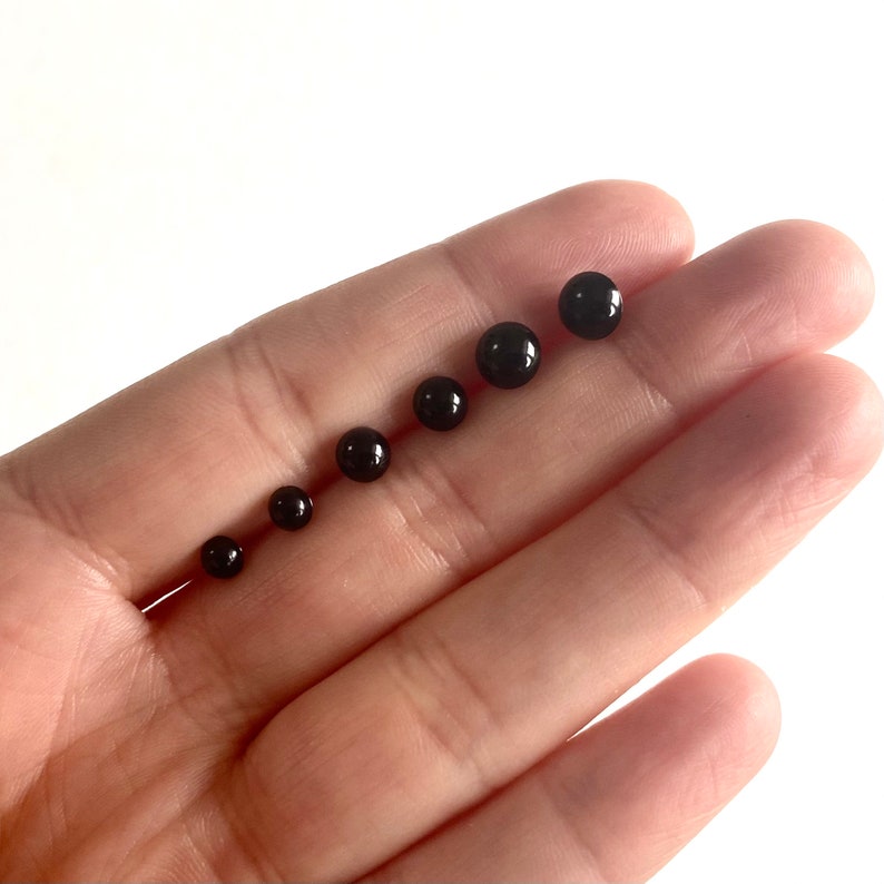 May include: A set of six black round beads of varying sizes, arranged in order from smallest to largest, on a hand.
