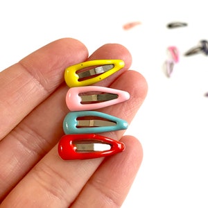 Può includere: Un set di cinque mini mollette per capelli colorate. Le mollette sono in tonalità di giallo, rosa, blu e rosso. Sono realizzate in metallo e hanno una finitura lucida.