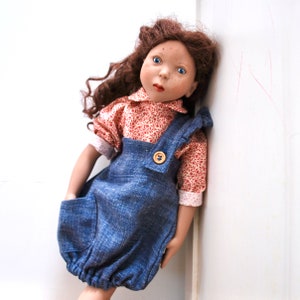 May include: A porcelain doll with brown curly hair wearing a red and white floral print shirt and blue denim overalls with a single button.