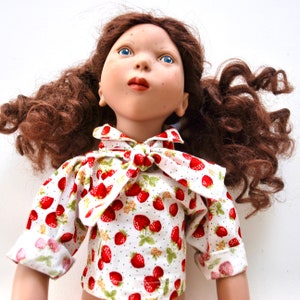 May include: A porcelain doll with long, curly brown hair wearing a white blouse with a red strawberry print and a bow tie.