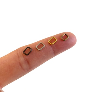 May include: Four small, rectangular metal rings in black, silver, gold, and bronze hues. The open-frame shapes are displayed on a fingertip against a white background. These are likely jewelry findings or components for crafting.