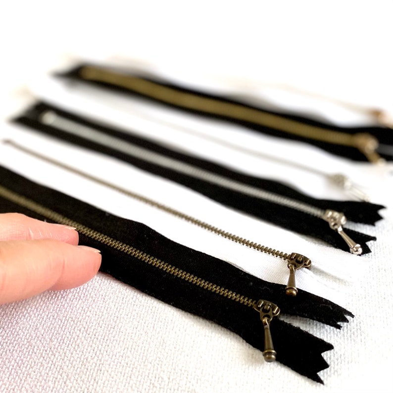 May include: A close-up of several black and white zippers with brass pulls. The zippers are laid out on a white surface.