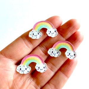 Pode incluir: Três pingentes de arco-íris de resina com arco-íris de cor com uma forma de nuvem em cada extremidade. Cada nuvem tem um rosto sorridente desenhado.