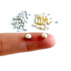 Könnte beinhalten: Zwei perlmuttartige Perlen, eine mit einer silbernen Fassung und die andere mit einer goldenen Fassung, werden auf einem Finger gezeigt.