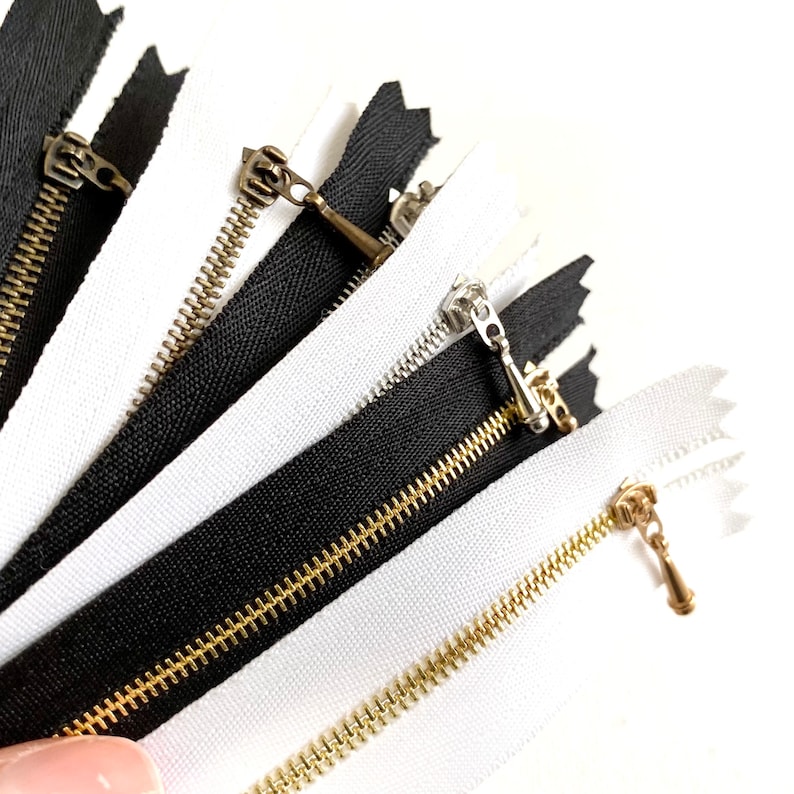 May include: A close-up of a variety of black and white zipper tapes with gold and silver zipper pulls. The zipper tapes are arranged in a fan shape.