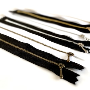 May include: A close-up of several black and white zippers with brass pulls. The zippers are arranged in a row, with the black zippers on top of the white zippers.