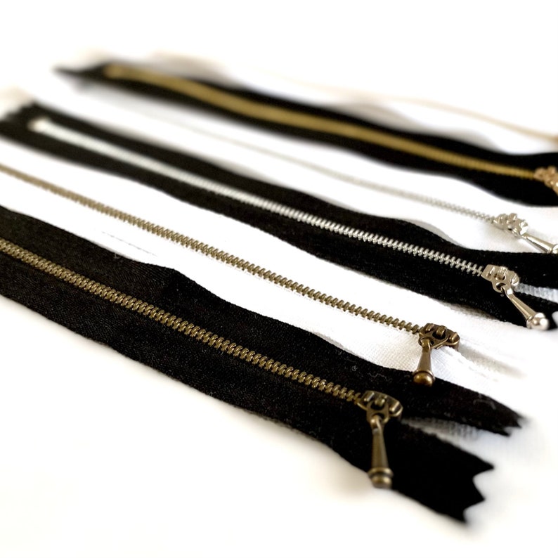 May include: A close-up of several black and white zippers with brass pulls. The zippers are arranged in a row, with the black zippers on top of the white zippers.