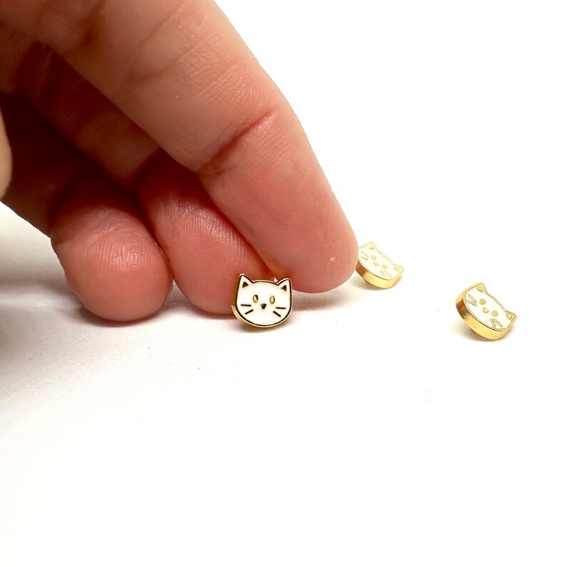 May include: Three gold-colored cat charms with white enamel. The charms are small and feature a simple cat face design.