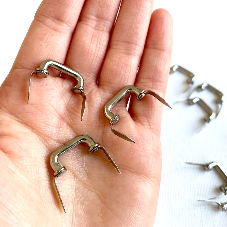 K&ouml;nnte beinhalten: Nahaufnahme einer Hand, die mehrere kleine silberne Metallgriffe h&auml;lt. Die Griffe haben eine D-Form und sind zum Anbringen an Taschen oder anderen Gegenst&auml;nden gedacht.