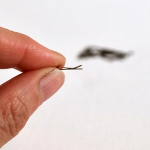 May include: A silver bobby pin, also known as a hair clip, is held between two fingers. The pin is about 2 inches long and has a slight bend at the end.