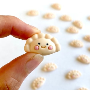 Könnte beinhalten: Eine Sammlung kleiner, weißer Keramik-Knödel mit fröhlichen Gesichtern. Jeder Knödel hat rosa Wangen und einen lächelnden Mund.