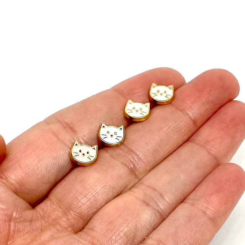 May include: Four gold-colored cat face charms with white enamel. The charms are small and have a simple design.