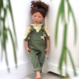 May include: A doll with brown hair wearing a yellow and white patterned shirt and green overalls. The doll is standing near a white wall.