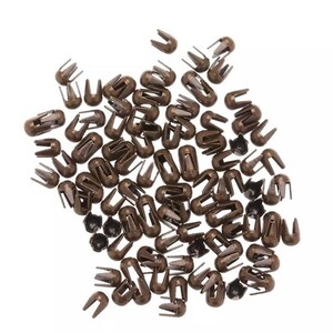 May include: A large pile of small, bronze-colored metal rivets with a rounded head and two prongs. The rivets are used for attaching fabric or leather.