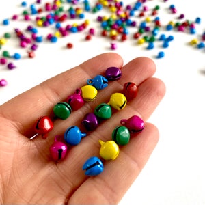 Könnte beinhalten: Eine Sammlung bunter Metall-Glöckchen mit einer silbernen Schlaufe zum Aufhängen. Die Glöckchen sind in verschiedenen Farben, darunter Rot, Blau, Grün, Gelb und Lila.