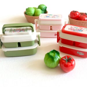 Peut inclure: Plusieurs boîtes à lunch empilables et colorées avec des poignées et des illustrations de dessins animés sur les couvercles. Les boîtes à lunch sont vertes, roses et rouges. Des tomates et de la laitue décoratives en céramique sont au premier plan et en arrière-plan.
