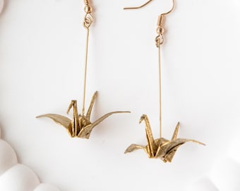 May include: A pair of gold-colored drop earrings featuring a metallic origami crane pendant suspended from a thin vertical bar and a standard fishhook ear wire. The crane design is detailed with folded wing and tail shapes in a metallic finish.