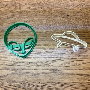 May include: Two cookie cutters, one shaped like an alien head and the other shaped like a flying saucer. The alien cookie cutter is green and the flying saucer cookie cutter is white.