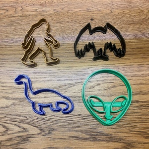 May include: Four cookie cutters in different shapes: a brown Bigfoot, a black bat, a blue dinosaur, and a green alien head.