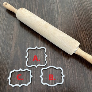 May include: A wooden rolling pin with a light brown handle and a white label on the side. Three white cookie cutters are on a dark brown wooden surface. The cookie cutters are shaped like ornate frames and have the letters A, B, and C inside.