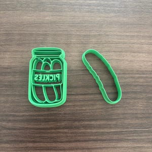 May include: A bright green plastic cookie cutter set. One cutter is shaped like a jar with the word "PICKLES" in reverse. The other cutter is shaped like a pickle.