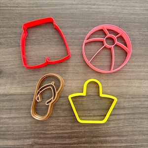 May include: Four cookie cutters in the shapes of summer items. There is a red pair of shorts, a pink beach ball, a brown flip-flop, and a yellow tote bag. The cutters are made of plastic and are on a wooden surface.