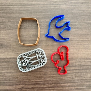 May include: Four plastic cookie cutters in the shapes of a barrel, a fish, a can of sardines, and a rooster. The barrel is tan, the fish is blue, the sardines are white and gray, and the rooster is red. The cutters are on a wooden surface.