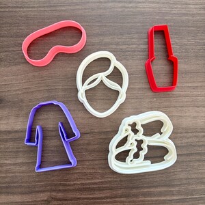 May include: Five plastic cookie cutters in various shapes. Includes a pink eye mask, a white face, a red nail polish bottle, a purple robe, and a white pair of sandals. The cutters are arranged on a wooden surface.
