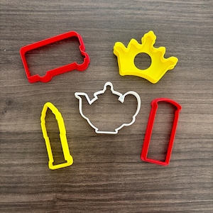 May include: A collection of colorful cookie cutters in various shapes, including a red bus, a yellow crown, a white teapot, a yellow bullet, and a red rectangle. These are likely used for baking and creating themed cookies.