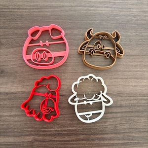 May include: Four plastic cookie cutters in the shapes of farm animals. The pig is pink, the cow is tan, the chicken is red, and the sheep is white. The cutters are on a wooden surface.