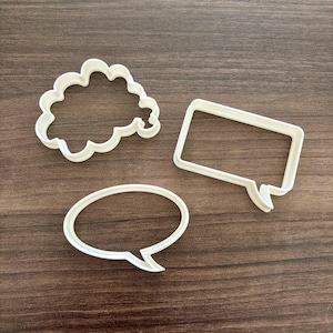 May include: Three white plastic cookie cutters in the shapes of speech bubbles. One is a cloud shape, another is a rectangle with a tail, and the third is an oval with a tail.