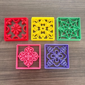 May include: Five colorful square cookie cutters in red, yellow, green, pink, and purple. Each cutter features a unique floral or geometric design within a square frame. The cutters are made of plastic and are arranged on a wooden surface.