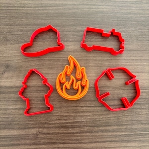 May include: A collection of red and orange cookie cutters in the shape of a firefighter's helmet, fire truck, fire hydrant, fire, and a Maltese cross. The cookie cutters are made of plastic and are arranged on a wooden surface.