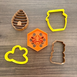 May include: Five cookie cutters in the shape of a beehive, honey pot, cloud, honeycomb with a bee, and a stack of honey. The cutters are in shades of yellow, orange, and brown.