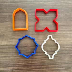 May include: Four colorful cookie cutters in orange, red, blue, and white. The shapes include an arched window, a geometric X, a starburst, and a teardrop design. These are ideal for baking cookies.