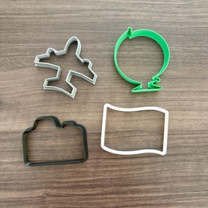 May include: Four cookie cutters in the shapes of an airplane, a globe, a camera, and a flag. The airplane is light gray, the globe is green, the camera is black, and the flag is white. The cutters are arranged on a wooden surface.